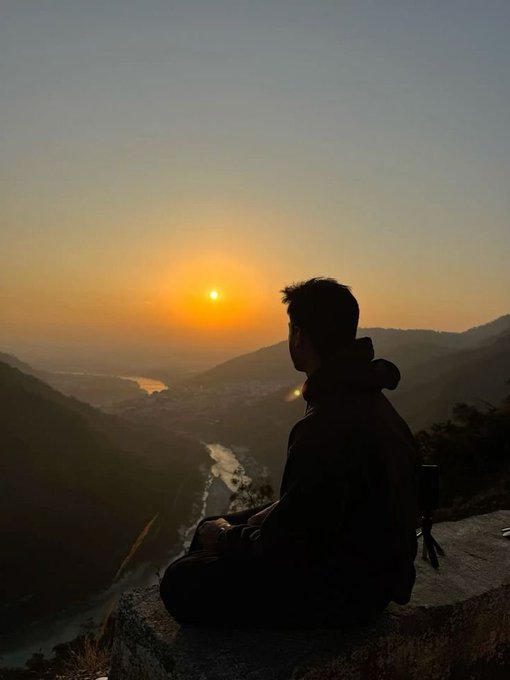 A person in a dark jacket sits silhouetted on a cliff, watching the sunset over a valley with a winding river.