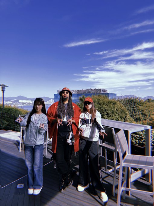 Three people pose together on a rooftop terrace with mountains and a stadium in the background on a sunny day.