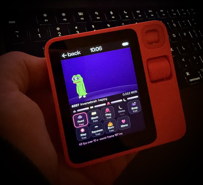 A person holds a red handheld device displaying a 'Inverebab happy' tamagotchi-style game with options like 'Feed', 'Wash', and 'Sleep' on a purple screen.