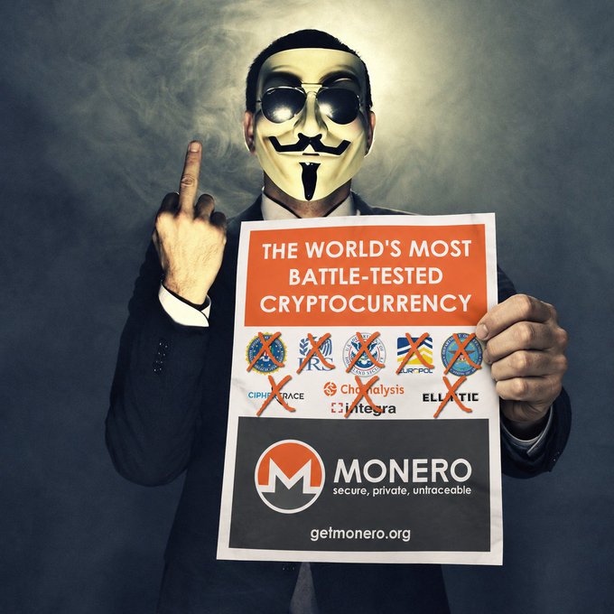 A person wearing a Guy Fawkes mask and sunglasses holds up a poster reading "THE WORLD'S MOST BATTLE-TESTED CRYPTOCURRENCY" promoting Monero with the tagline "getmonero.org", showing logos of competing blockchain analytics firms crossed out.