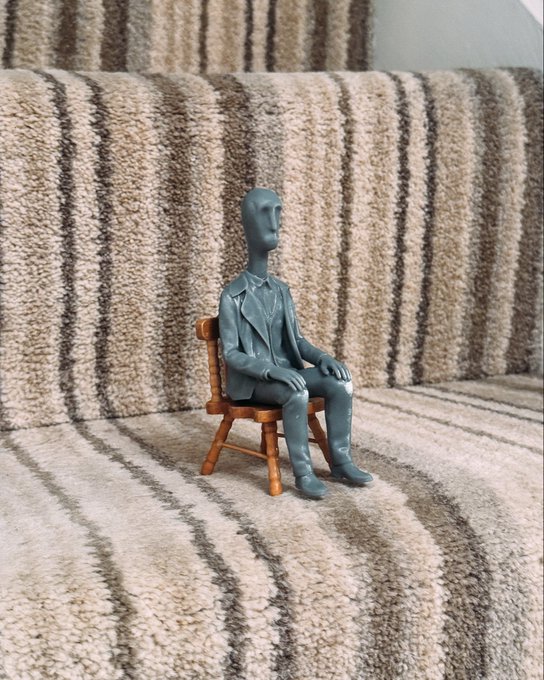 A small gray sculpted figure of a suited man sits on a miniature wooden chair atop a striped brown and cream carpet.