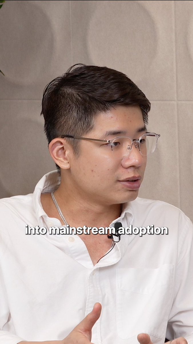 A young man with short black hair and clear glasses, wearing a white collared shirt and a lavalier microphone, speaking in an interview setting. The subtitle 'into mainstream adoption' is visible across his chest.