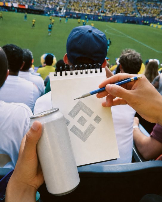 A hand sketches the Binance logo on a notepad while holding an aluminum can, with a soccer match unfolding in the stadium stands behind them—perfectly capturing the "Matchday view, Binance edition" vibe of blending crypto culture with live sports.