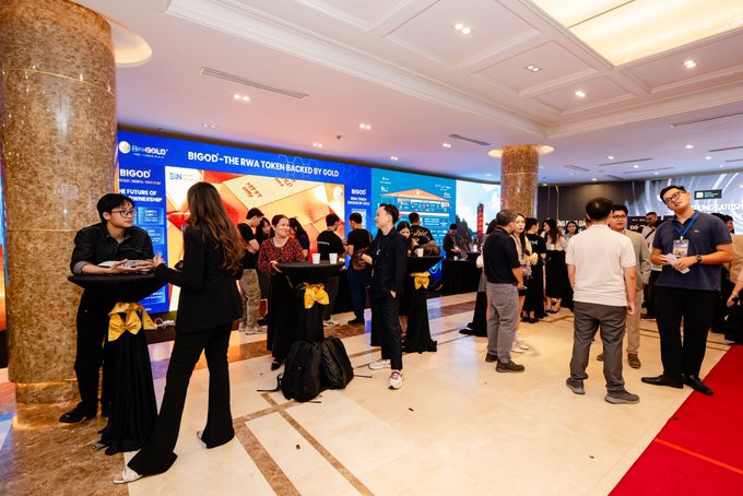 An indoor corporate event or networking gathering takes place in an upscale venue with marble floors and columns, where attendees in business attire mingle near cocktail tables, with a large screen displaying 'BiGOD - THE RWA TOKEN BACKED BY GOLD' and 'FEATURES OF BiGOD' in the background.