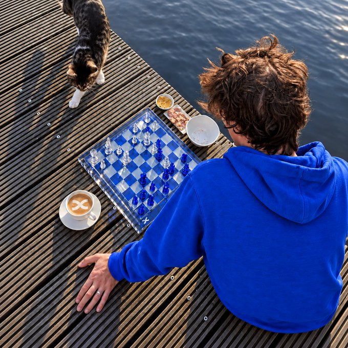 A boy in a blue hoodie sits on a wooden dock beside a chess board, a coffee cup, and a curious tabby cat looking down at the water. The image pairs with "gm to everyone playing the long game♟️" by capturing a quiet, strategic moment—chess symbolizing patience and foresight in a peaceful waterside setting.