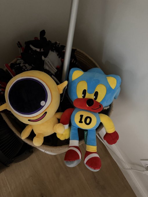 Two plush toys sit in a wicker basket, including a yellow Minion with one eye and a blue fox character wearing a red scarf and yellow shirt with the number '10'.