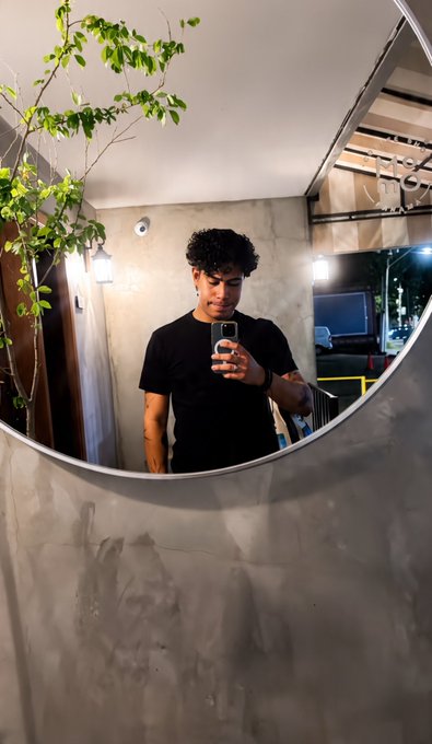 A young man with curly hair wearing a black t-shirt takes a mirror selfie with his phone in a uniquely designed space with a curved mirror and a hanging plant.