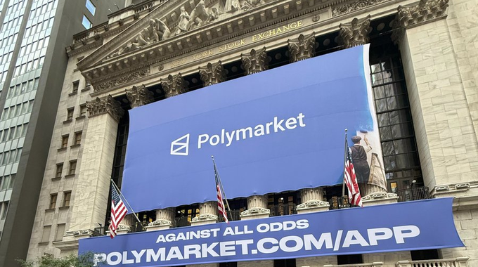 A banner promoting Polymarket hangs above the New York Stock Exchange, displaying the company's logo and "AGAINST ALL ODDS" tagline alongside the URL "POLYMARKET.COM/APP." The image ties to the post by visually celebrating Polymarket's major financial milestone—a $1 billion raise at a $21 billion valuation—through a prominent display at one of the world's most iconic financial landmarks.