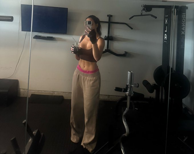 A mirror selfie in a home gym showing a woman in a brown sports bra, beige sweatpants, and workout gloves, holding a glass and her phone, with an Ironax cable machine visible behind her.