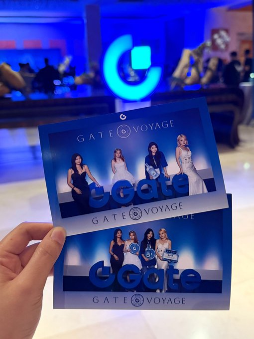 A hand holds two promotional cards for "GATE VOYAGE" featuring four female performers in elegant dresses, with a large illuminated "G" logo in the blue-lit background.