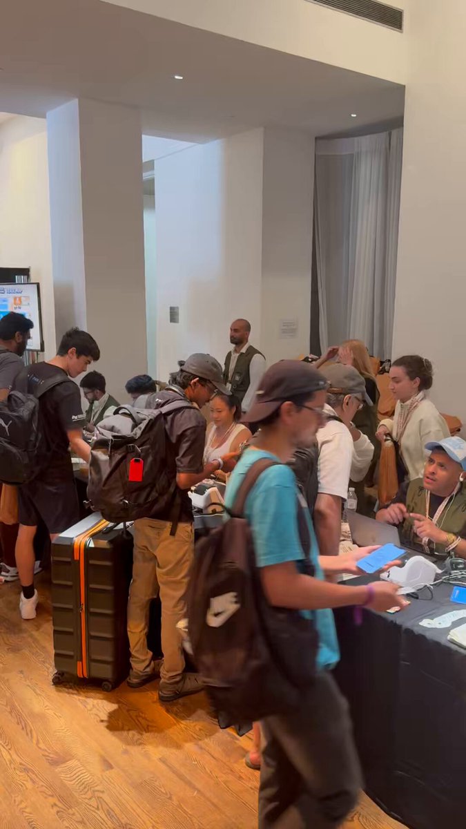 The image shows a group of people standing around, some with luggage such as backpacks and suitcases. They appear to be waiting for something or someone, possibly at an event like a conference or gathering. The room has white walls and wooden floors, giving it a clean and organized appearance. A TV is mounted on the wall in one corner of the room.