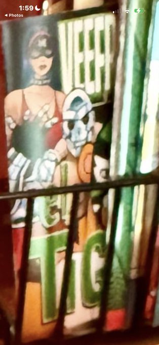 A vertically-held photo showing a comic book or VHS cover featuring a female superhero in a mask and a masked male character, with partial green text reading 'WEEP' and 'TIC' visible.
