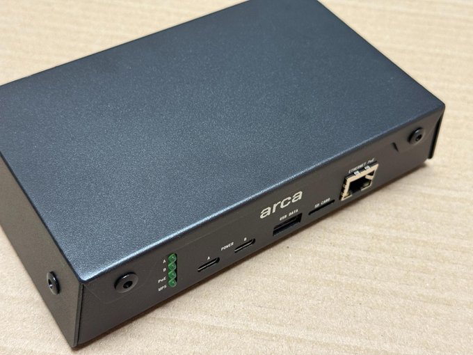 A black 'arca' branded mini PC or networking device sitting on a wooden surface, featuring ports for HDMI, USB, SIM, and Ethernet.