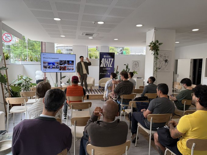 The image shows a group of people sitting in chairs around a table, watching a man giving a presentation using a large screen TV as a projection device. The room appears to be a conference or meeting space where attendees are engaged in listening to the speaker. The atmosphere seems to be professional and focused on the topic being discussed.