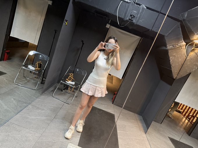 The image features a woman taking a selfie with her cell phone while standing next to two chairs. The chairs are positioned behind her, one of which has a "@binancezh" logo on it. The background includes a mirror that reflects the room's interior and a black door in the background.