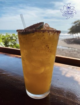 A tall glass of "The Stacy Maracuya Mocktail" — a passion fruit beverage with a chili-salt rim — sits on a wooden bar overlooking the beach, matching the post's tribute to its namesake namesake on the Bitcoin Country beach bar menu.