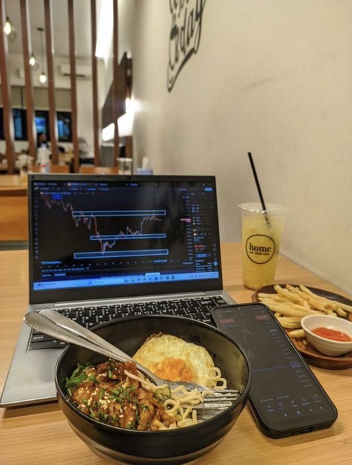 A workspace scene featuring a laptop displaying trading charts, a bowl of noodles topped with a fried egg, french fries with ketchup, a drink in a 'home' branded cup, and a smartphone showing trading data.