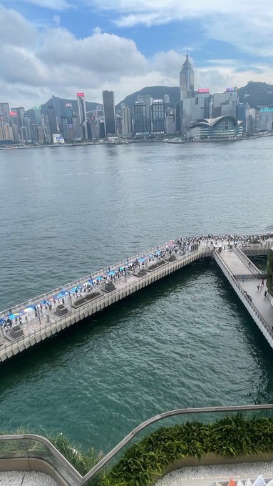 An elevated view of Hong Kong harbor shows a long pier crowded with people, the city skyline with skyscrapers in the distance, and the Hong Kong Convention and Exhibition Centre visible across the water.
