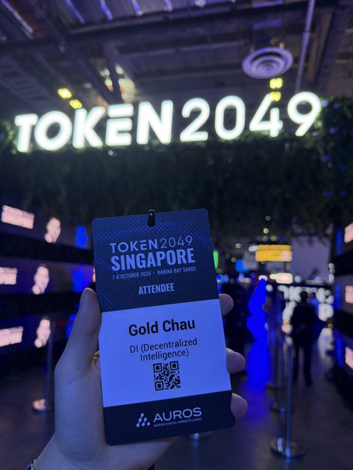 A hand holds an attendee badge for TOKEN2049 Singapore (7-8 October 2026, Marina Bay Sands) in front of a large illuminated 'TOKEN2049' sign. The badge displays the name 'Gold Chau' from 'DI (Decentralized Intelligence)' and is branded by AUROS.