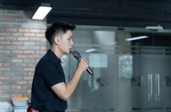 A young man in a black t-shirt holding a microphone, speaking or singing in what appears to be a warehouse or studio setting with exposed brick walls.