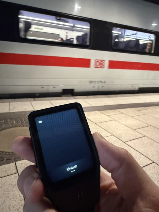A hand holding an iPhone displaying the 'Unlock' screen on a train platform, with a Deutsche Bahn (DB) train passing by in the background.