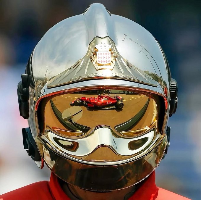 Close-up of a person wearing a chrome racing helmet with a Monaco coat of arms crest and a mirrored gold visor that reflects a red Formula 1 race car.