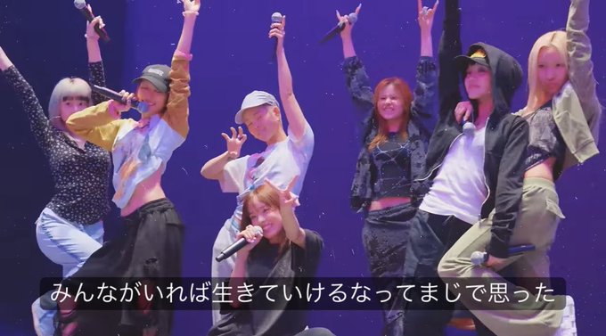 A group of performers on stage holding microphones with their arms raised, with Japanese subtitles reading 'みんながいれば生きていけるなってまじで思った' displayed at the bottom.