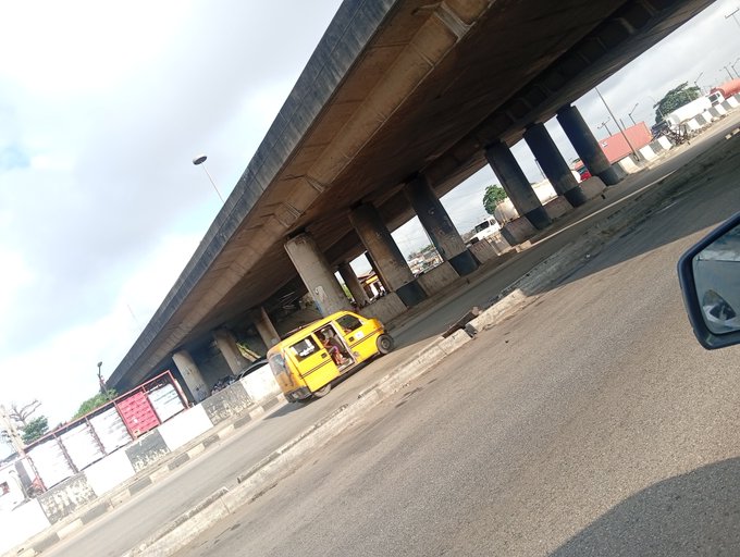 A yellow taxi drives on a road beneath a large elevated highway supported by concrete pillars.