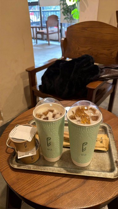 Two green beverages topped with ice and cream sit on a metal tray on a wooden table, branded with the 'OE' logo, alongside a receipt and small cups.