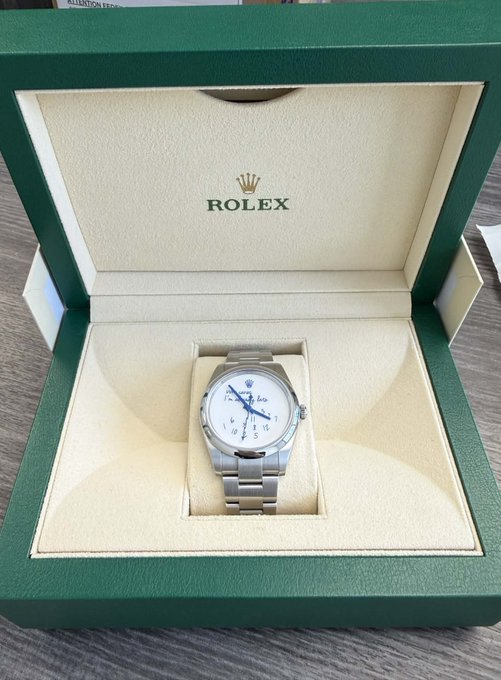 A silver Rolex watch with a blue dial sits inside a green presentation box with the 'ROLEX' crown logo embossed on the cream-colored interior lining.