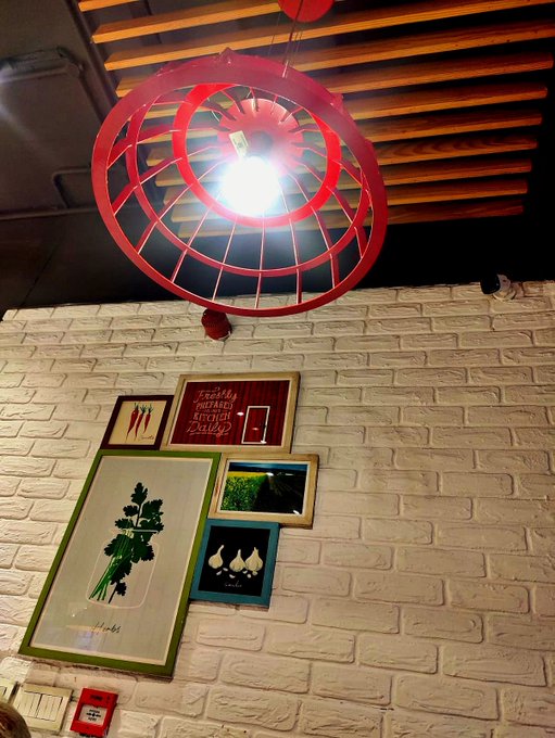 A red circular pendant light hangs from a wooden ceiling above a white brick wall decorated with framed botanical prints and artwork, including one labeled "KITCHEN GALLERY".