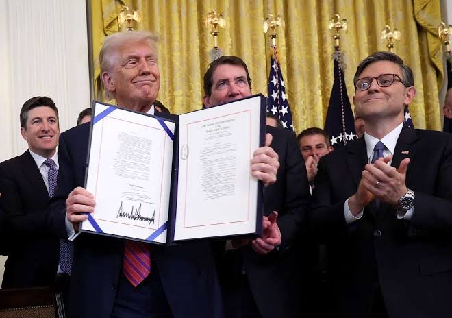 Former President Donald Trump proudly holds up a signed document in the White House, flanked by smiling officials, including one in glasses applauding on the right. The caption reflects a ceremonial signing event, with the post connecting this imagery to anticipation around the upcoming CLARITY Act vote and potential benefits for XRP.