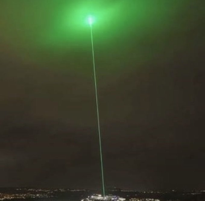 Nighttime cityscape featuring a tall tower shooting a bright green laser beam into the sky.