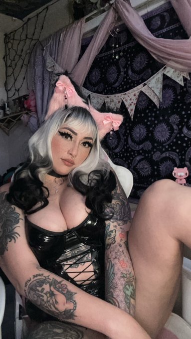 A selfie of a woman with silver hair, pink cat ears, and extensive tattoos on her arms, wearing a black PVC corset-style top in a decorated room.