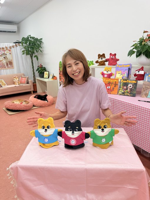A smiling woman in a pink shirt sits at a table with three plush dog toys wearing colored shirts, surrounded by Fantaulmi merchandise and a cat resting nearby.
