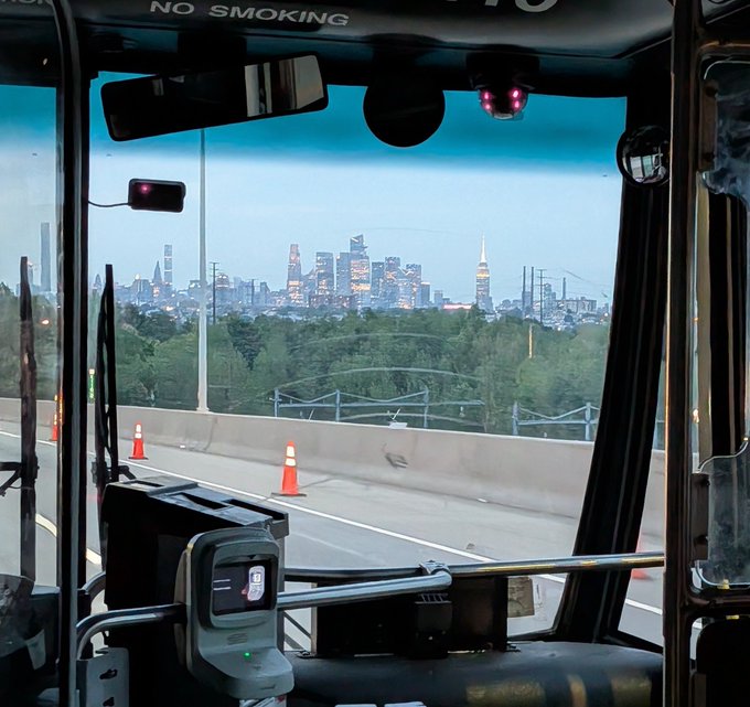 A view from inside a bus looking out at the New York City skyline across a highway, with traffic cones visible on the road and a 'NO SMOKING' sign at the top.