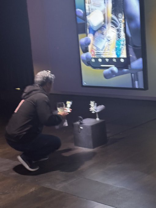 A person in dark clothing crouches down in front of a large screen displaying an image of a hand holding a smartphone, reaching toward some small objects on a pedestal.
