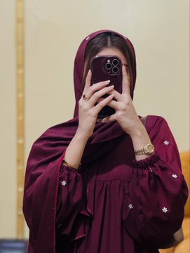 A mirror selfie of a woman wearing a maroon hijab and matching embroidered dress, holding a smartphone in front of her face.