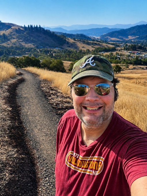 A smiling man in a camouflage "A" cap and maroon "California 1772" t-shirt takes a selfie on a gravel trail overlooking golden hills and a valley, embodying a cheerful "Good Morning" greeting to fellow greeters enjoying the scenic morning view.