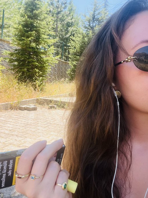 A close-up selfie of a woman wearing sunglasses and wired earphones, holding a tube of sunscreen, with multiple rings on her fingers against an outdoor background of trees and a stone wall.