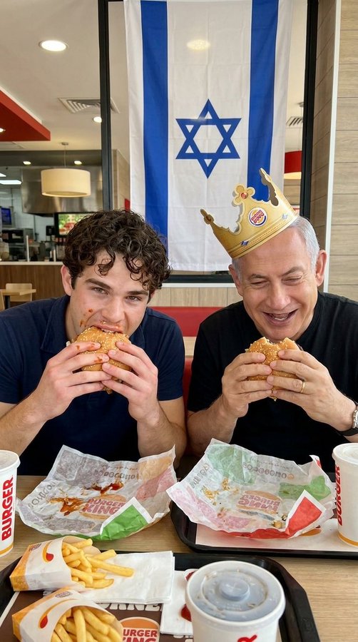 Two men eating Burger King Whoppers at a fast food restaurant, with one wearing a gold Burger King crown, seated beneath a large Israeli flag on the wall.