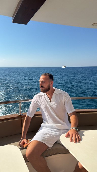 A bearded man in a white polo shirt and shorts relaxes on the deck of a yacht with the deep blue ocean stretching out behind him, perfectly capturing the essence of a luxurious "vibe check."
