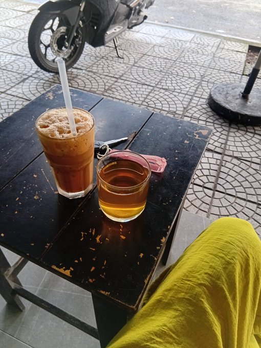 A casual outdoor café scene featuring a glass of iced coffee with a straw and a smaller glass of what appears to be tea or honey on a weathered black table, with a parked motorcycle and tiled sidewalk in the background.