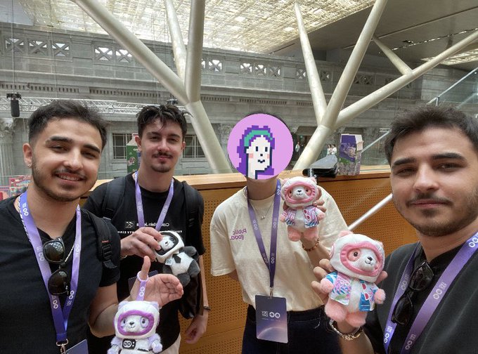 Four people pose for a photo, with three of them holding small plush toys; one person's face is obscured by a pink pixelated avatar and 'OO' lanyards are visible.