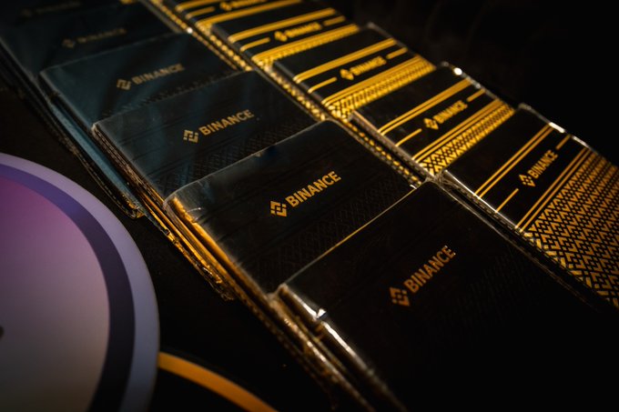 Stacks of black and gold cards or packets branded with the 'BINANCE' logo are arranged on a dark surface.
