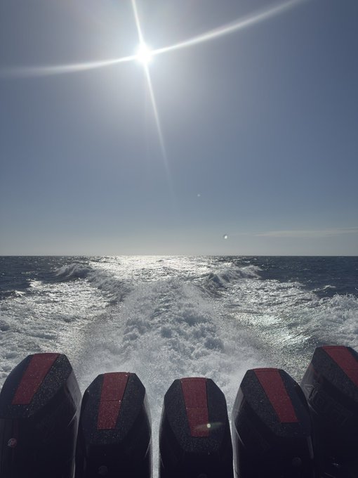 A view from the stern of a boat showing four outboard motors churning up white water as the vessel speeds across the sea under a bright sun.
