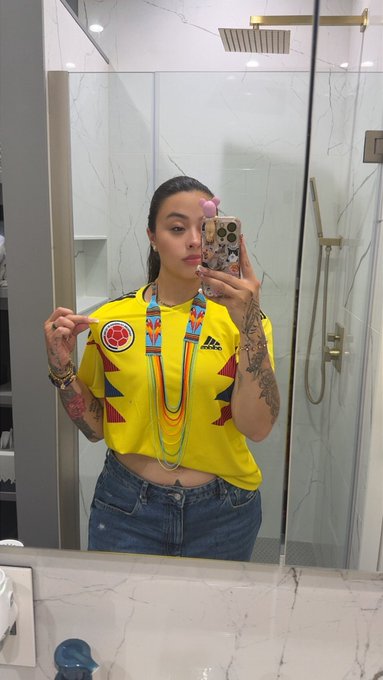 A mirror selfie of a tattooed woman in a bathroom wearing a yellow Adidas Colombia soccer jersey and blue jeans, pointing at the crest on her shirt.