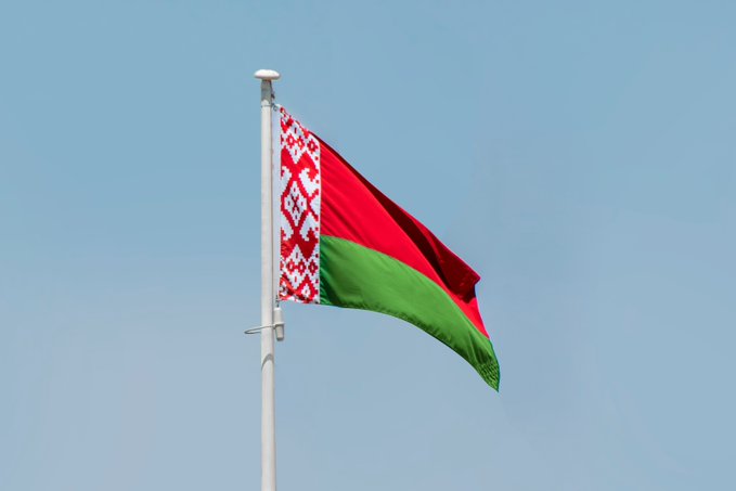 The national flag of Belarus, featuring its distinctive red and green stripes with a white ornamental pattern, waves against a clear blue sky on a flagpole. The image illustrates the post's news about the EU imposing restrictions on Belarusian nationals and residents from owning or managing MiCA-regulated crypto service providers starting August 25.