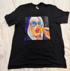 A black t-shirt laid out on a white surface, featuring a painted portrait of a person with a white head covering, blue eyeshadow, and bright red lips holding a yellow object.