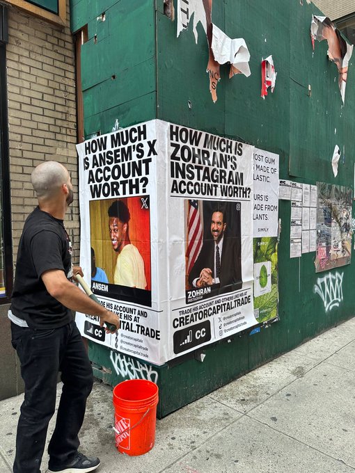 A man stands on a sidewalk next to a green wall displaying posters reading 'HOW MUCH IS ANSEM'S X ACCOUNT WORTH?' featuring Ansem, and 'HOW MUCH IS ZOHRAN'S INSTAGRAM ACCOUNT WORTH?' featuring Zohran Mamdani, with 'CREATORCAPITALTRADE' branding.